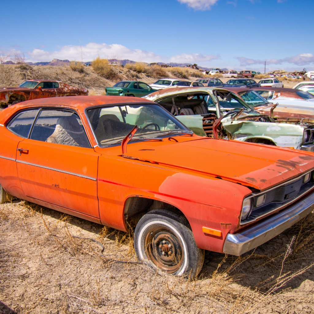 Rusty orange vintage car sitting in a desert junkyard surrounded by abandoned vehicles, illustrating a scrap car scenario.