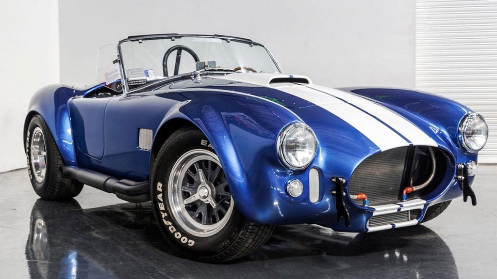 A classic blue Shelby Cobra with white racing stripes parked indoors on a glossy black floor. The open-top roadster features chrome accents, Goodyear racing tires, and a sleek, curvaceous body design.