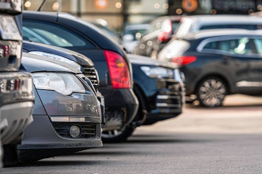 A close-up view of parked cars in a busy parking lot, with a blurred background showing a commercial or shopping area.