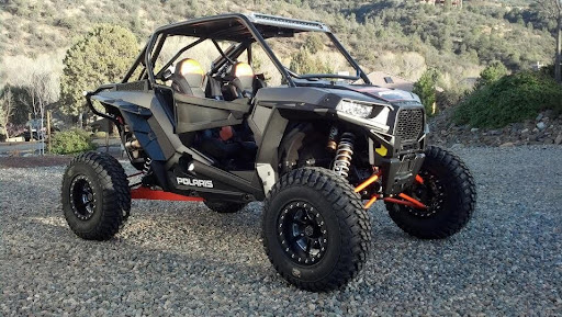 A rugged Polaris side-by-side (UTV) parked on a gravel surface with a scenic mountainous background, featuring large off-road tires, a reinforced roll cage, and an upgraded suspension system.