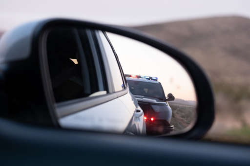 A close-up view of a car's side mirror reflecting a police vehicle with flashing lights during a traffic stop on a remote road with hills in the background.