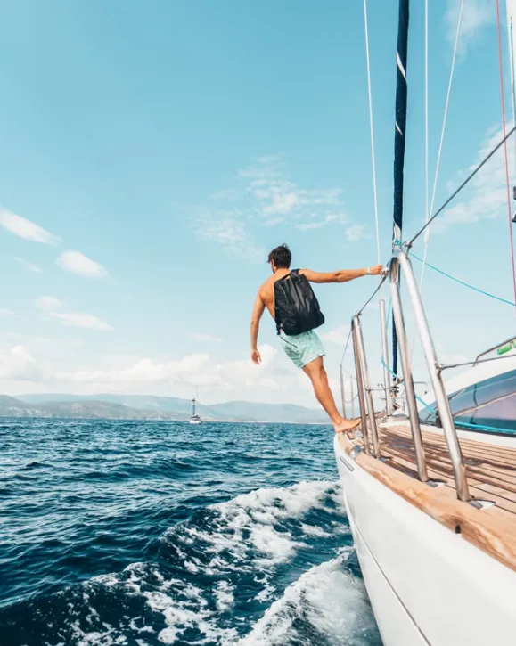man with a backpack stands on the edge of a sailboat cruising through vibrant blue waters - Dirt Legal A man with a backpack stands on the edge of a sailboat cruising through vibrant blue waters under a clear sky.