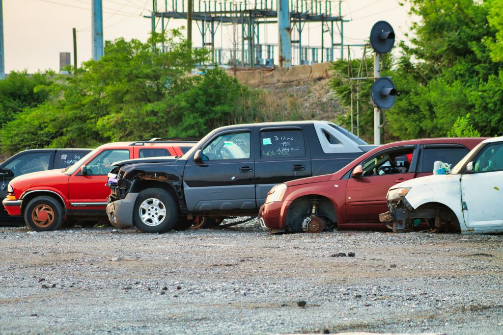 The image shows a row of damaged and abandoned vehicles parked in a gravel lot, with missing wheels, broken parts, and visible signs of wear, likely at a salvage yard or impound lot.