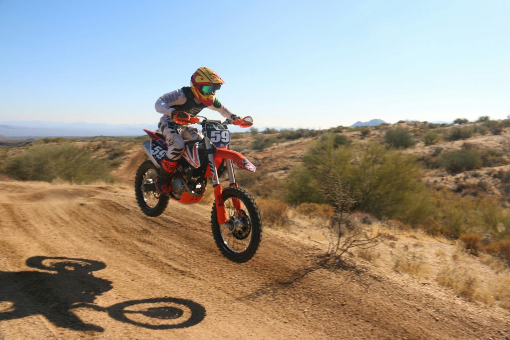 A motocross rider wearing a helmet and goggles launches off a dirt jump in the desert, mid-air on a number 59 bike under a clear blue sky.