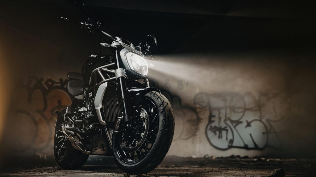 A black motorcycle sits in a dark graffiti-covered room with its headlight shining forward.