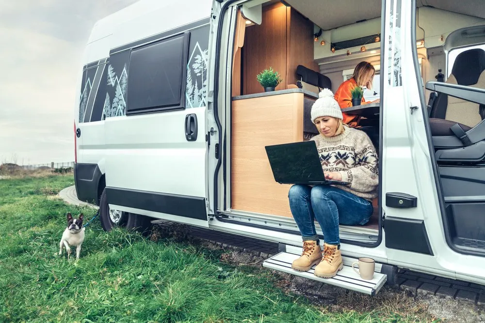 woman working on a laptop while sitting on the step of a camper van, with another person inside working at a table and a small dog on a leash nearby - Dirt Legal This image shows a woman working on a laptop while sitting on the step of a camper van, with another person inside working at a table and a small dog on a leash nearby, capturing a cozy and modern van life scene in a grassy outdoor setting.
