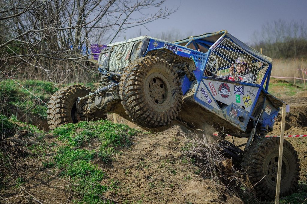 A rugged off-road vehicle covered in sponsor decals tackles a steep, muddy hill during an extreme off-roading event, with the helmeted driver focused on the climb.