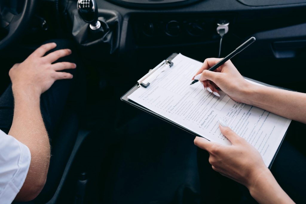 The image shows a person filling out a form on a clipboard inside a car, likely during a driving test or vehicle inspection.