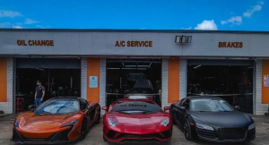 Three high-end sports cars—a McLaren, a Lamborghini, and an Audi R8—are parked in front of an auto repair shop offering oil changes, A/C service, and brake work, with a technician visible near the garage.