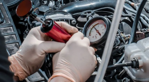 A technician wearing gloves is using a diagnostic pressure gauge and testing tool to inspect a vehicle's engine components up close.