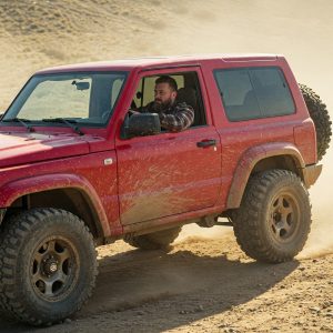 A bearded man in a plaid shirt grips the wheel of a mud‑splattered red off‑road SUV as it kicks up dust across a rugged dirt trail.