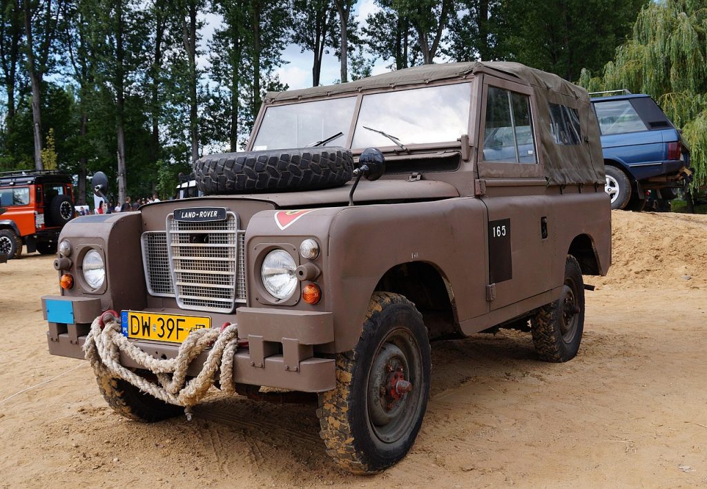 A classic Land Rover Defender, equipped for adventure with a rugged exterior and front-mounted gear, stands ready on a dirt trail surrounded by other off-road vehicles.