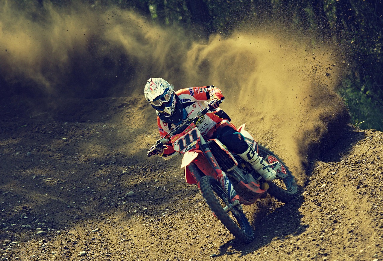 A motocross rider in full racing gear leans sharply into a turn on a dirt track, kicking up a large cloud of dust and debris behind the bike.
