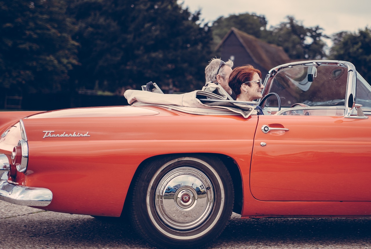 An older couple enjoys a sunny drive in their bright red vintage Ford Thunderbird convertible, proud to have secured its title and made it road legal.
