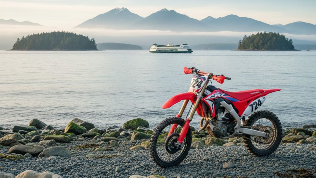 Dirt bike parked on a rocky beach overlooking Puget Sound in Washington, with evergreen islands, misty mountains, and a ferry boat in the distance.