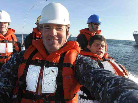 U.S. Navy Crew at Sea - Dirt Legal A group of U.S. Navy personnel wearing camouflage uniforms, life jackets, and safety helmets are riding in a small boat on the open water, smiling and taking a selfie during what appears to be a training or transport mission.