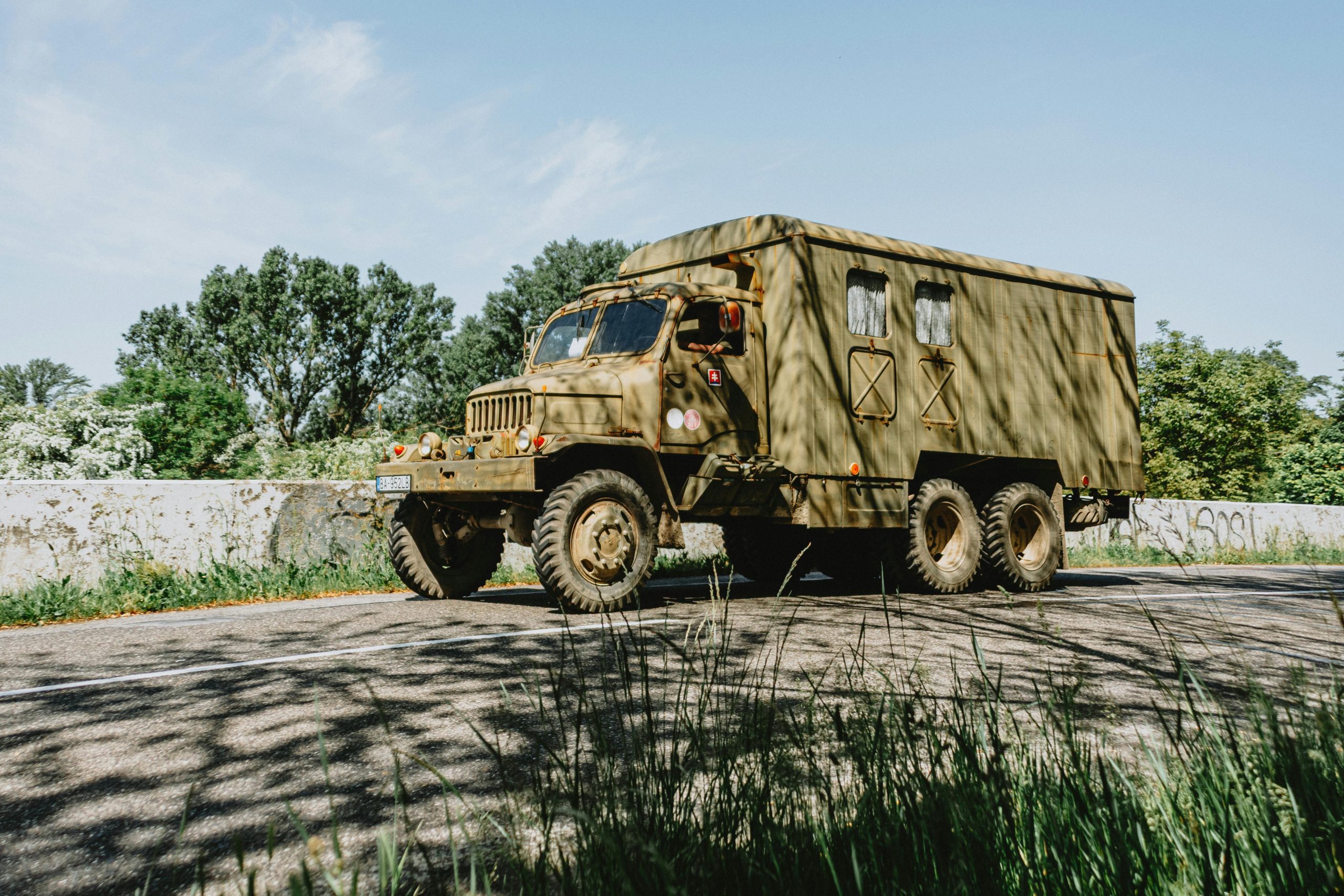 A vintage military cargo truck with a canvas-covered rear drives along a quiet road, surrounded by trees and greenery, showcasing its rugged, utilitarian design.