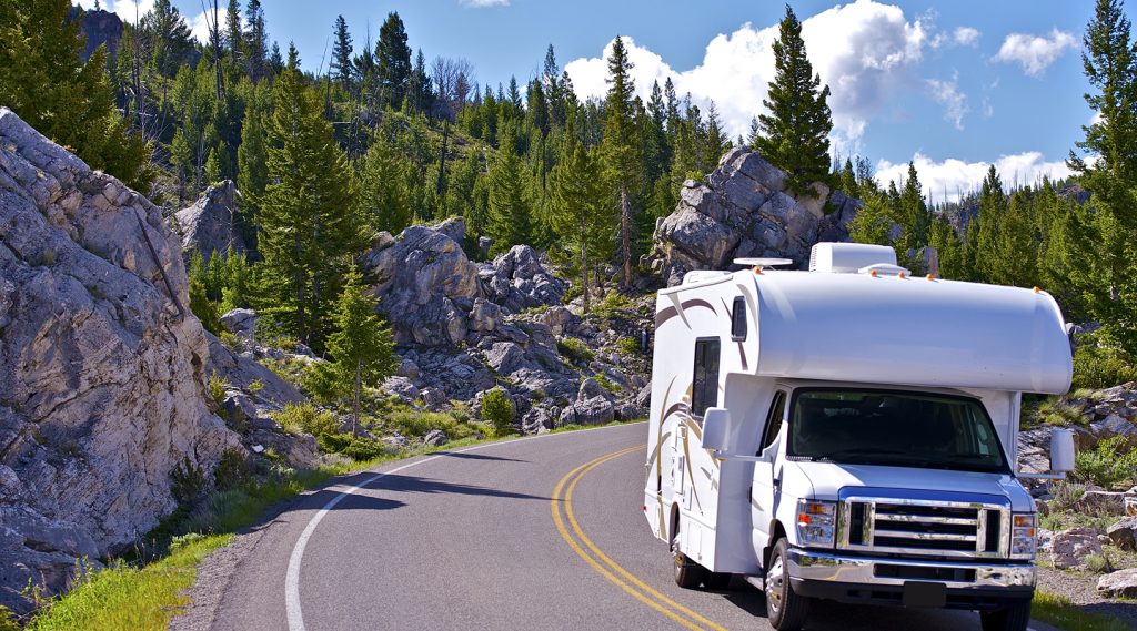 White Class C RV driving along a scenic mountain road surrounded by pine trees and rocky cliffs.