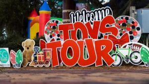 Toys Tour event sign with red letters, teddy bear, train, bike, Christmas lights, and bouncy castle in background