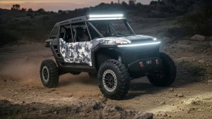 Toyota Scion 01 Hybrid SxS Off-Road Concept - SEMA 2025 - Dirt Legal Toyota Scion 01 Concept off-road vehicle with digital camo livery driving on a dirt trail at dusk, showcasing its LED light bar and powerful stance. This hybrid SxS debuted at SEMA 2025.