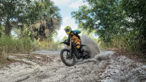 E-Moto rider on a performance-upgraded Sur Ron electric dirt bike performing an aggressive off-road wheelie and kicking up dirt, showcasing GritShift parts.
