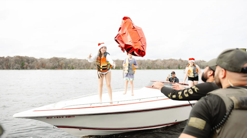People in Santa hats and life vests on a speedboat throwing a large red wrapped present into the water