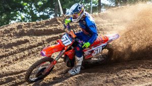 Supercross rider cornering fast on a KTM dirt bike through deep sand berm during a race, wearing a white helmet and blue/orange gear.