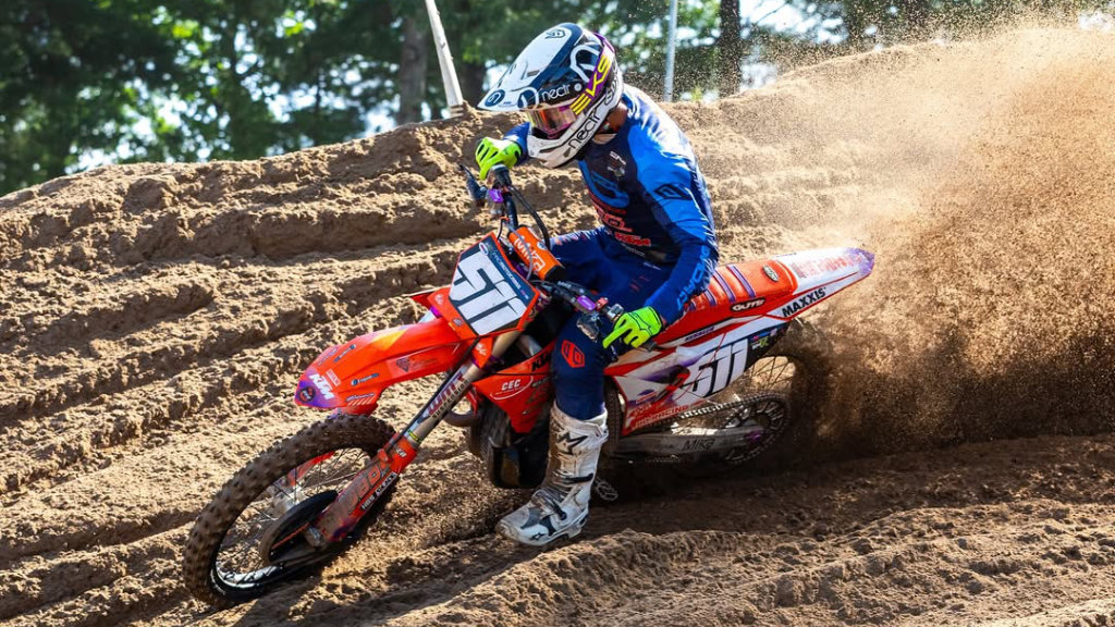 Supercross rider cornering fast on a KTM dirt bike through deep sand berm during a race, wearing a white helmet and blue/orange gear.