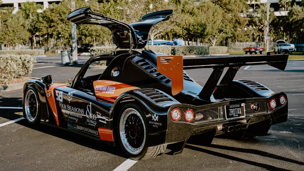 Rear view of a black Ultima GTR race car with gull-wing doors open, featuring a large carbon fiber spoiler and a Montana ZROTAX license plate.