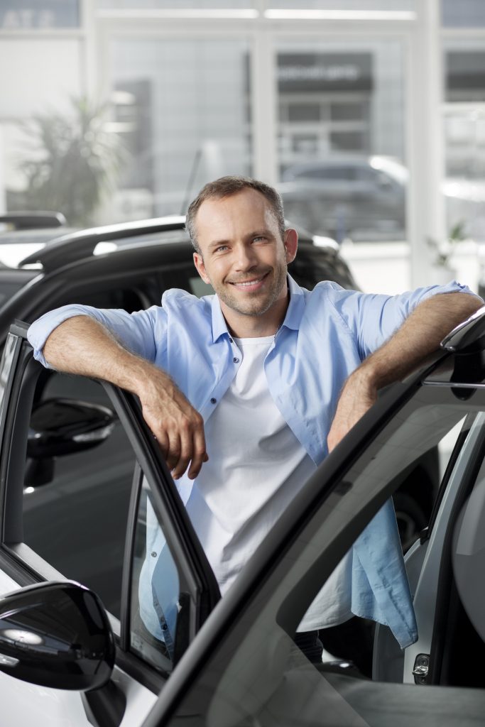 A smiling man leans confidently against his open car door in a bright showroom, looking like a happy driver registered vehicle forming a momntana llc.