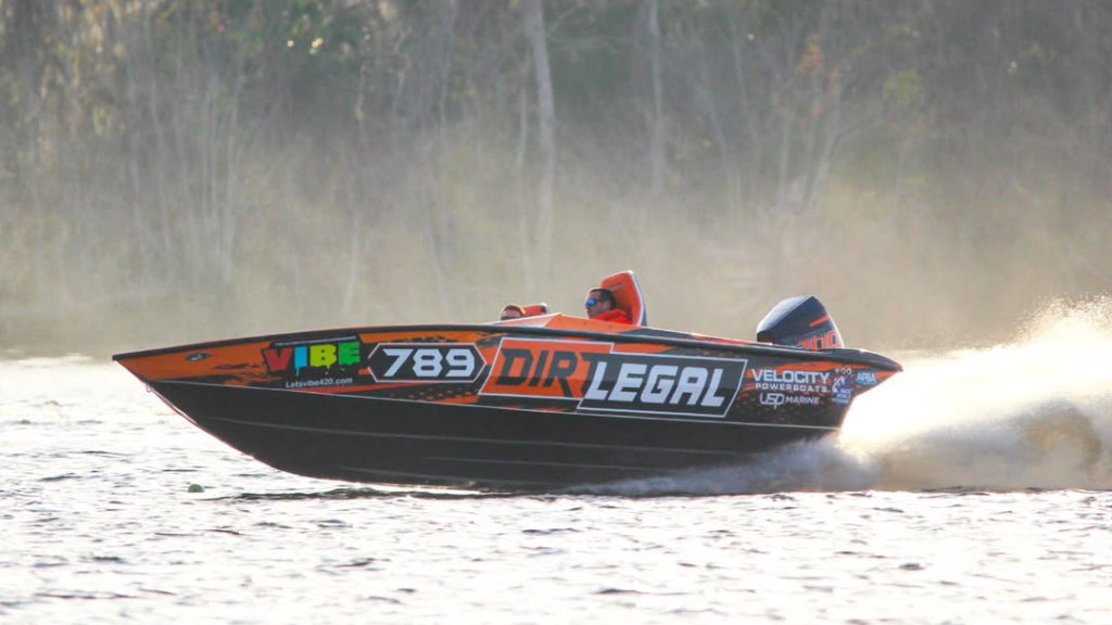 A black and orange high-performance Velocity powerboat with Dirt Legal branding cuts through the water, creating a large white spray at high speed.