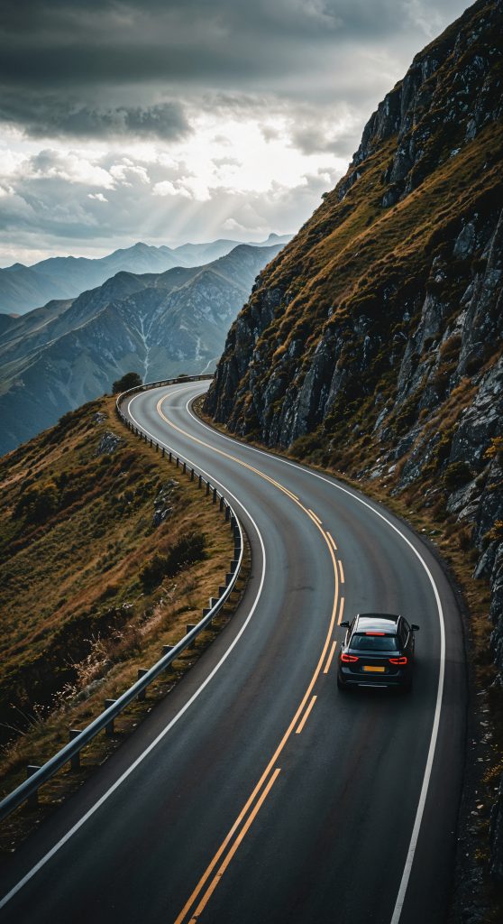 A lone car drives along a winding mountain road beneath dramatic clouds with sunlight streaming over distant peaks.