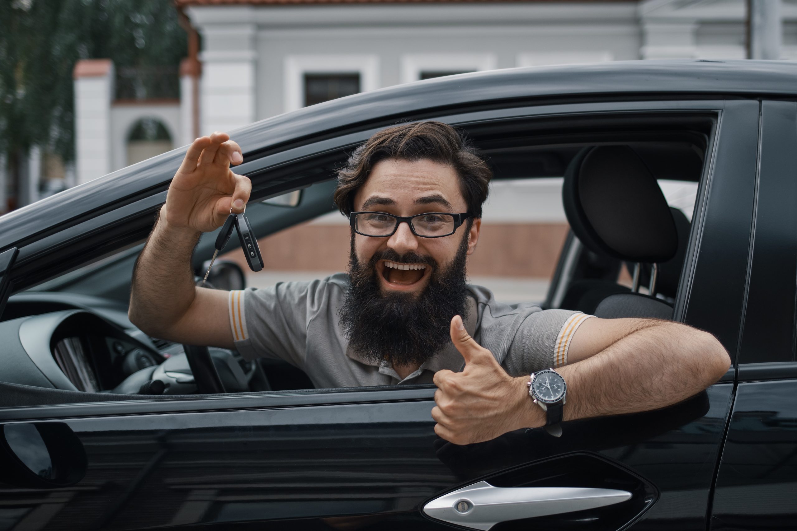 A smiling man sits in his car holding up the keys and giving a thumbs-up, representing someone who has successfully formed a Montana LLC and registered their vehicle with ease.