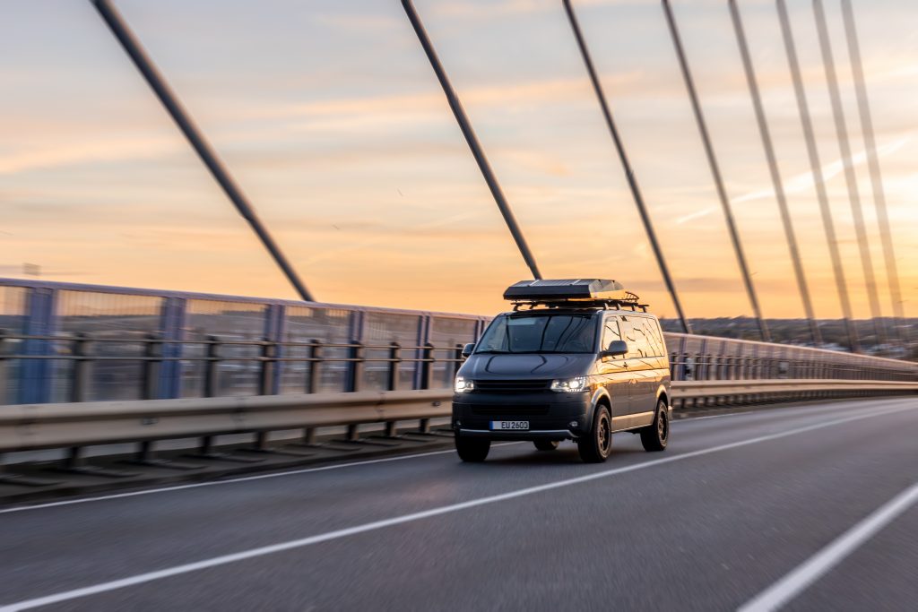 Dark camper van with rooftop cargo driving across a modern bridge during sunset.