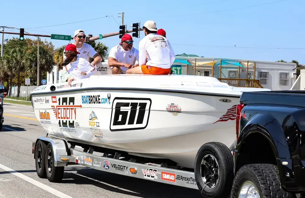 Powerboat with racing number 611 on a trailer being towed by a pickup truck, with several people sitting on the boat during a street boat parade