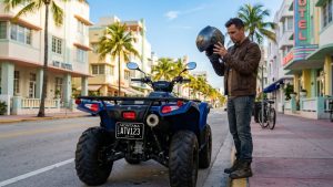 A rider putting on a helmet next to a street-legal ATV with a Montana license plate parked on a busy street in Miami Beach.
