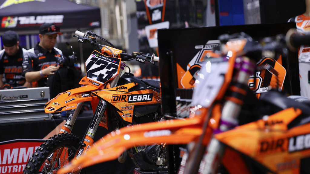 Multiple orange KTM race bikes with Dirt Legal graphics parked in the team pits with mechanics and toolboxes in the background.
