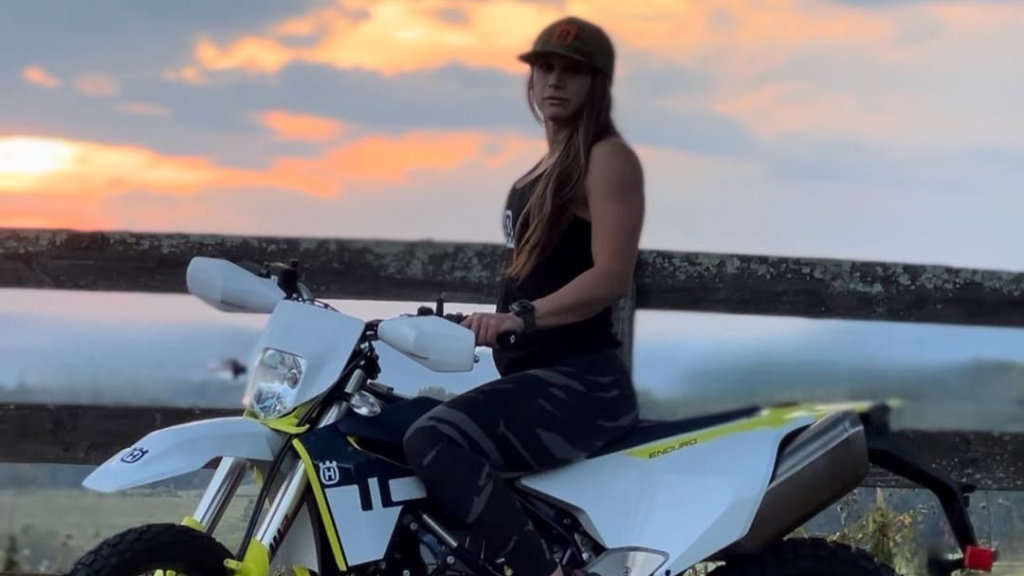 A woman in a hat and riding gear sitting on a white Husqvarna enduro motorcycle with a vibrant orange and blue sunset in the background.