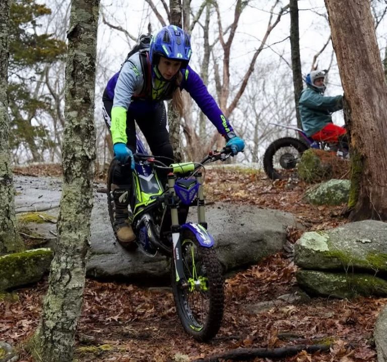 A rider wearing blue and purple gear skillfully maneuvering a trials dirt bike down a rocky embankment in a wooded area.