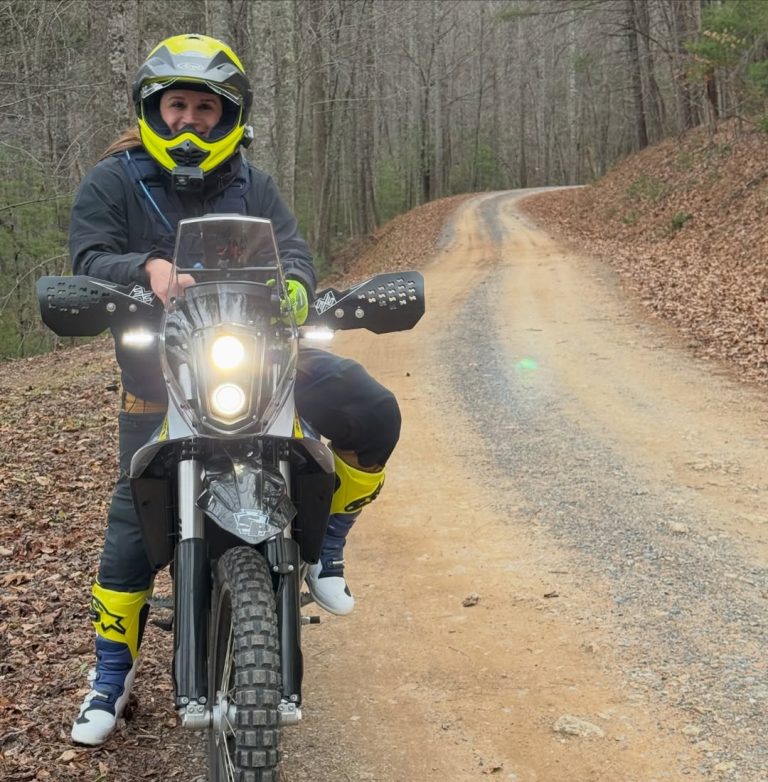A female rider in full gear and a yellow helmet sitting on a Husqvarna adventure motorcycle on a gravel trail surrounded by trees.