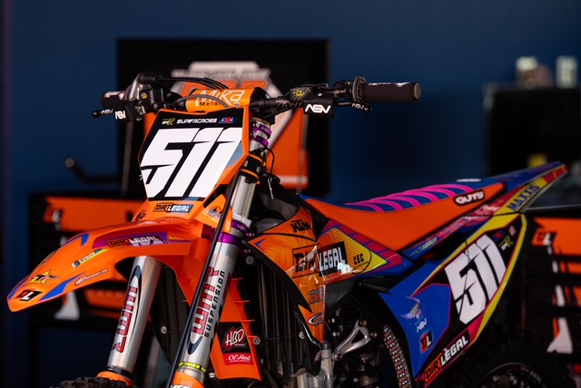 A detailed front-profile shot of a Dirt Legal DBD Racing WMR KTM dirt bike featuring custom HBD graphics, a GUTS Racing seat cover, and the number 511 plate in the team pits.