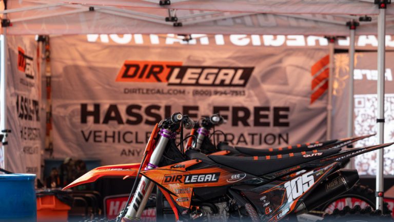 The Dirt Legal DBDRacing pit setup featuring a row of KTM race bikes under a branded tent. A large banner in the background promotes hassle-free vehicle registration services.