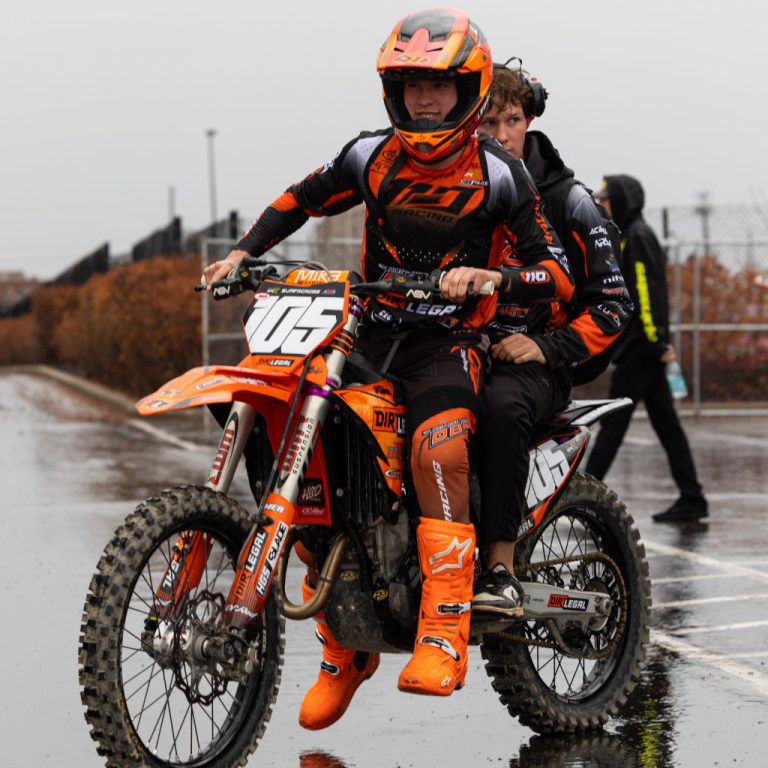 Marcus Phelps riding his orange and black KTM 250 through a wet Indianapolis parking lot with a team member on the back. Both are geared up in Dirt Legal DBDRacing colors on a rainy race day.