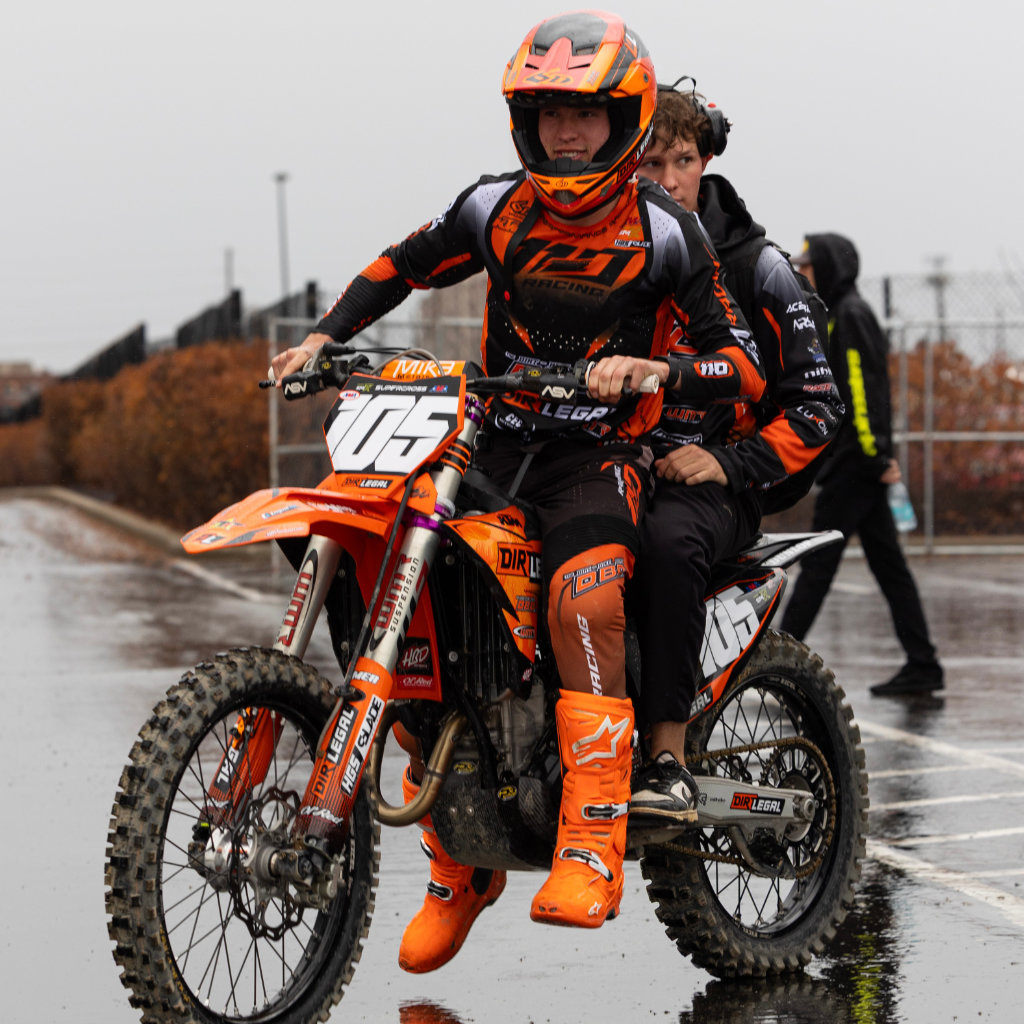 Marcus Phelps riding his orange and black KTM 250 through a wet Indianapolis parking lot with a team member on the back. Both are geared up in Dirt Legal DBDRacing colors on a rainy race day.