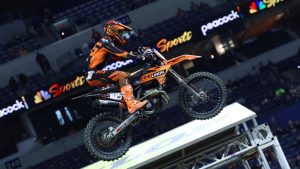Marcus Phelps on his #105 KTM mid-air over a tabletop jump at the Indianapolis Supercross. The bike features full Dirt Legal branding against the backdrop of the stadium seating and Peacock Sports banners.