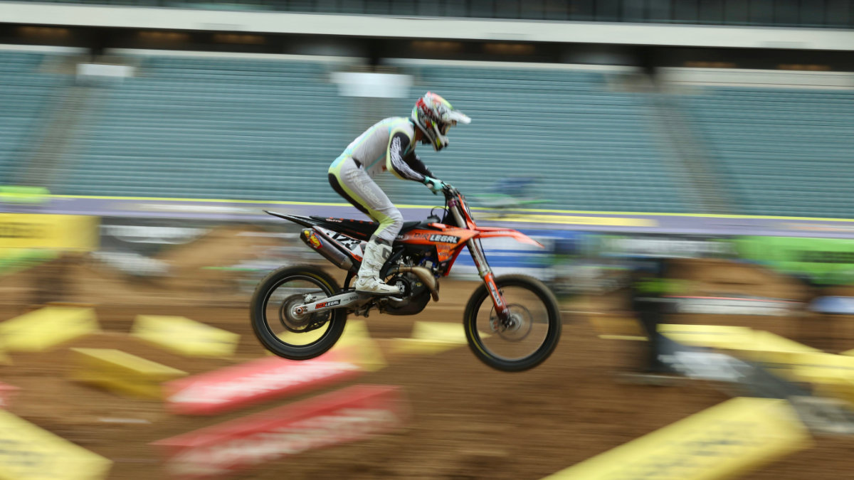A professional motocross rider in white gear with orange accents launches a Dirt Legal-branded KTM bike into the air during a stadium race.