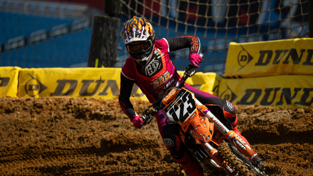 Anthony Rodriguez wearing a pink, purple, and black jersey and a flame-patterned helmet leans his orange KTM bike sharply through a deep dirt corner. He is racing on a track lined with yellow Dunlop barriers.