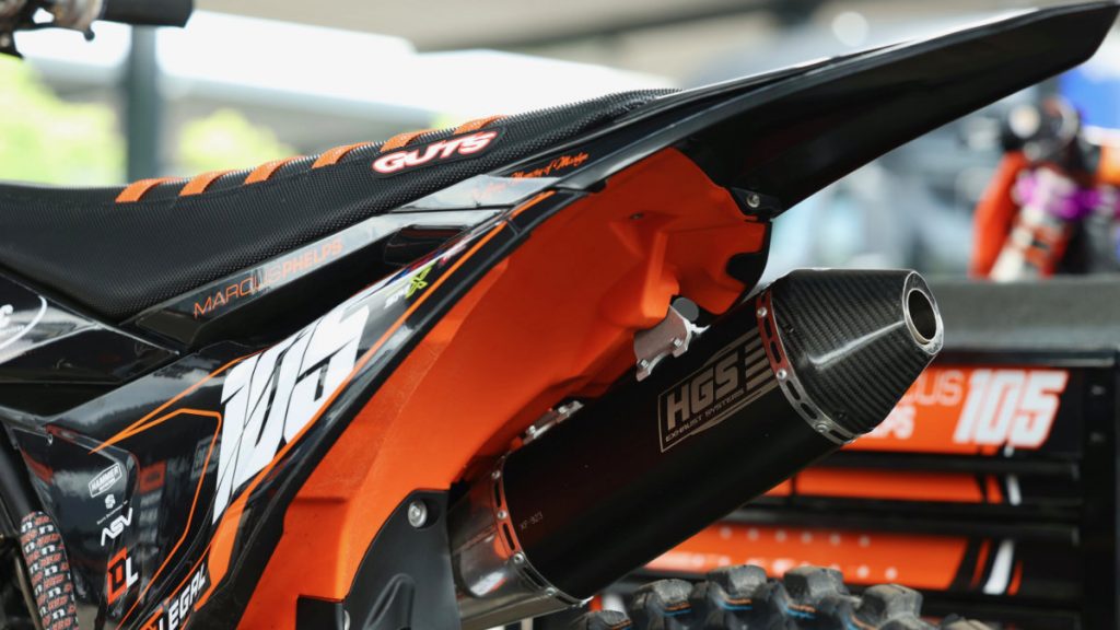 A close-up view of the rear of a custom Dirt Legal-sponsored KTM race bike, showing the black GUTS racing seat and the HGS exhaust system.