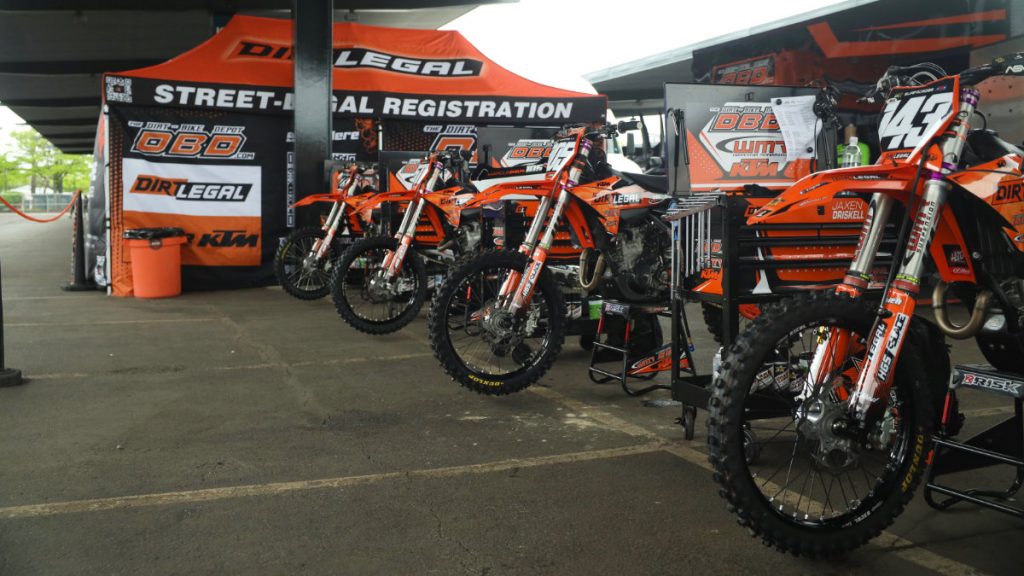 A long line of custom KTM motocross bikes stands ready under a Dirt Legal "Street-Legal Registration" team pit tent in a stadium paddock.
