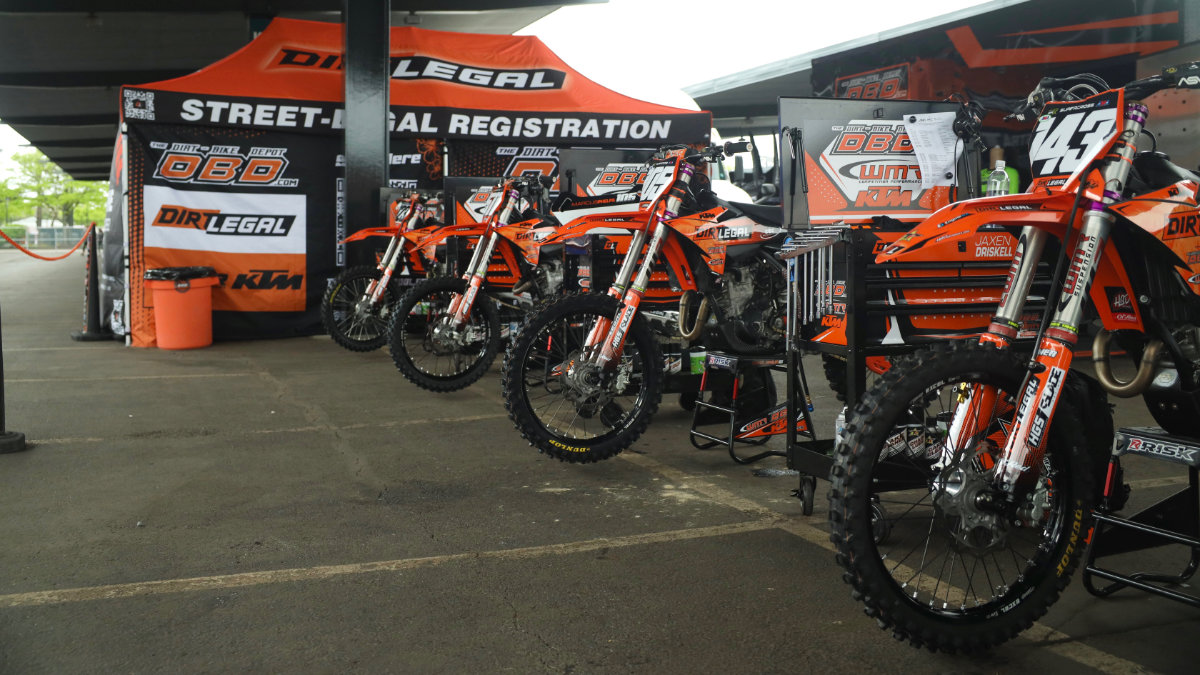 A long line of custom KTM motocross bikes stands ready under a Dirt Legal "Street-Legal Registration" team pit tent in a stadium paddock.